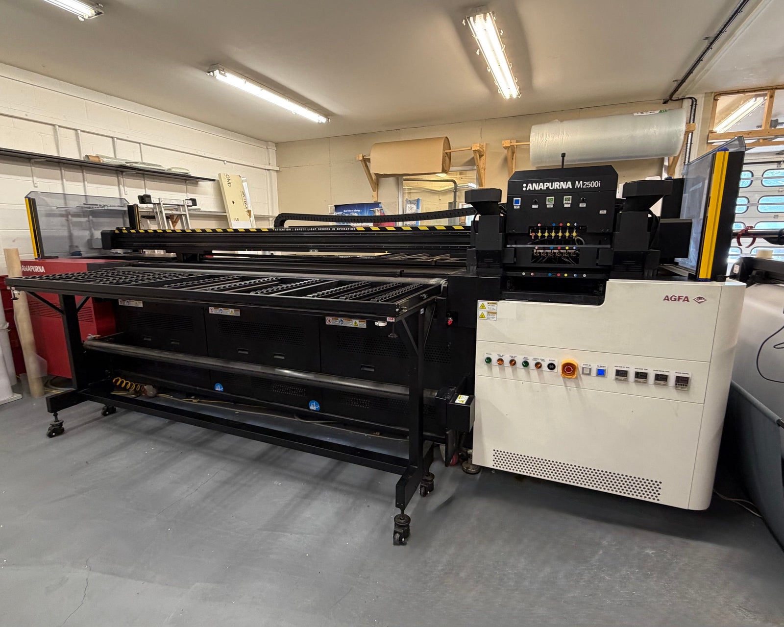 A large industrial flatbed UV printing machine, specifically a LANJIMA MV2000 with an AGFA scanner bed, photographed inside a workshop or garage environment with concrete floors and fluorescent lighting.