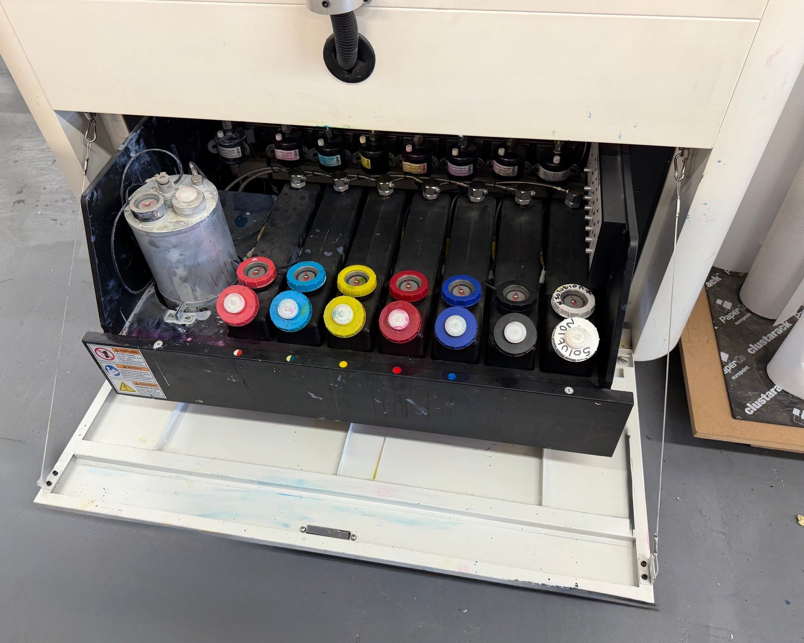 Open drawer of a desktop inkjet printer showing six black ink tanks with red, cyan, yellow, blue, and black ink cartridges, plus a gray reservoir tank.