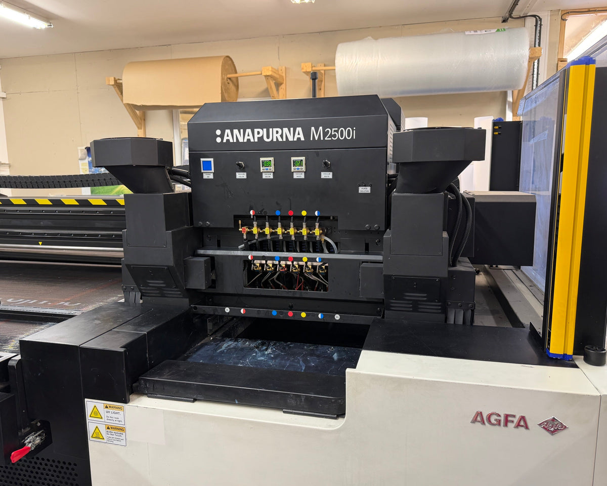 An industrial AGFA Anapurna M25001 UV printer shown in a workshop setting, featuring a black and white chassis with yellow safety accents and a digital control screen on the right.