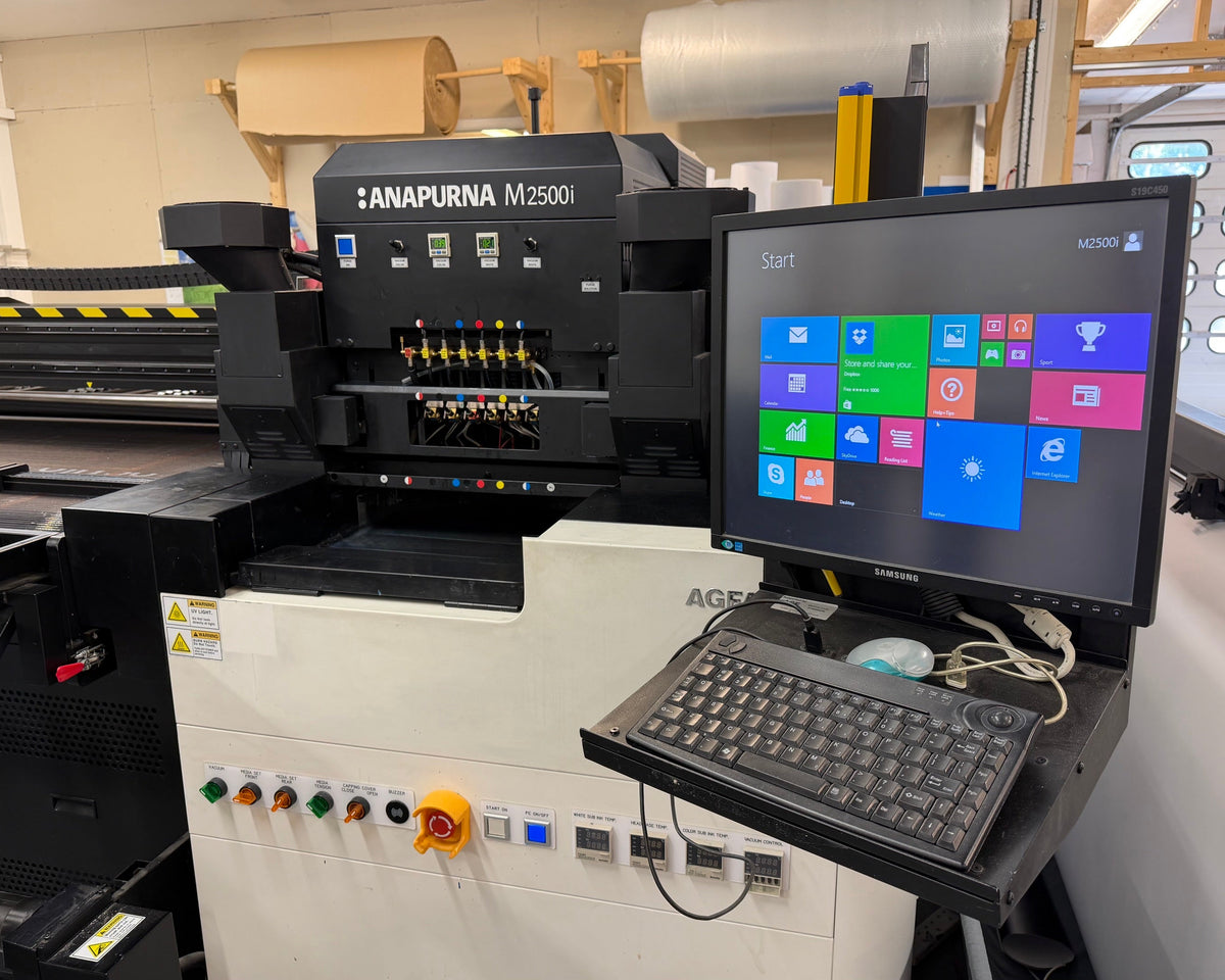 An industrial flatbed inkjet printing machine labeled 'ANAPURNA M2500i' with an attached computer monitor, keyboard, and control panel in a workshop setting.