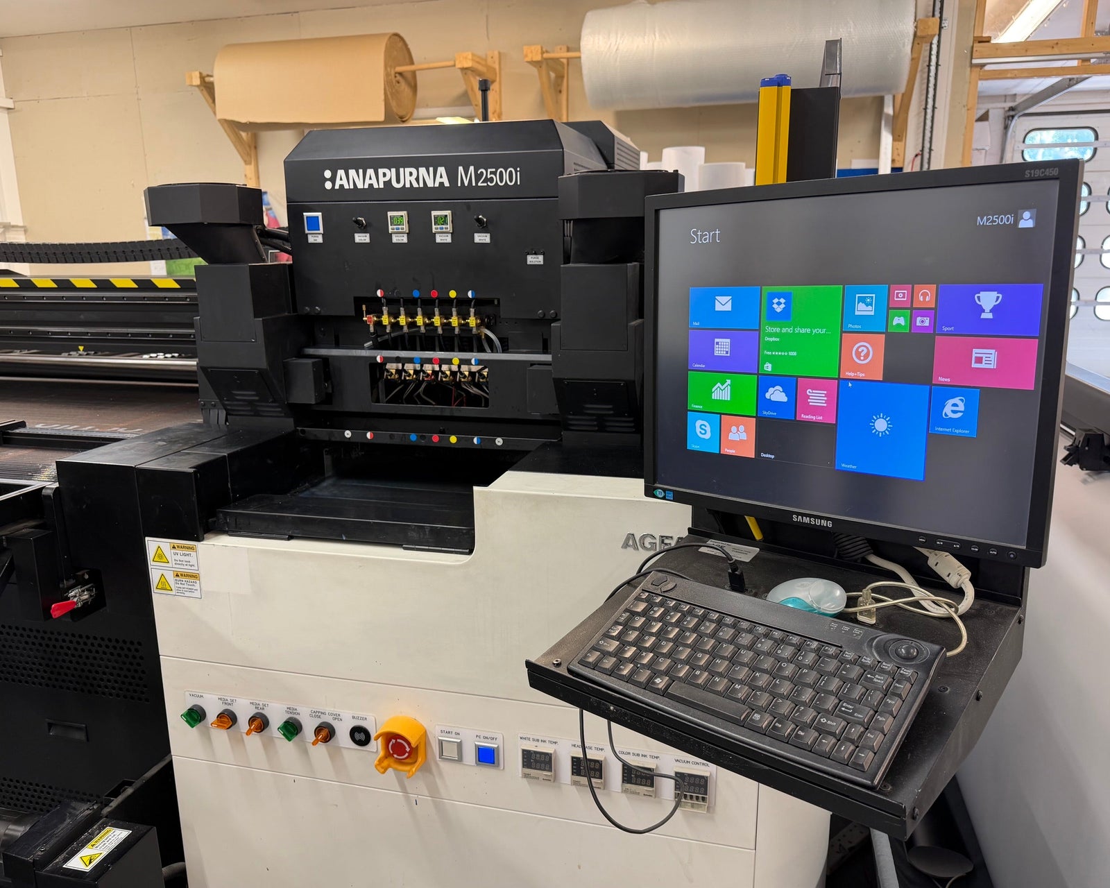 An industrial flatbed inkjet printing machine labeled 'ANAPURNA M2500i' with an attached computer monitor, keyboard, and control panel in a workshop setting.