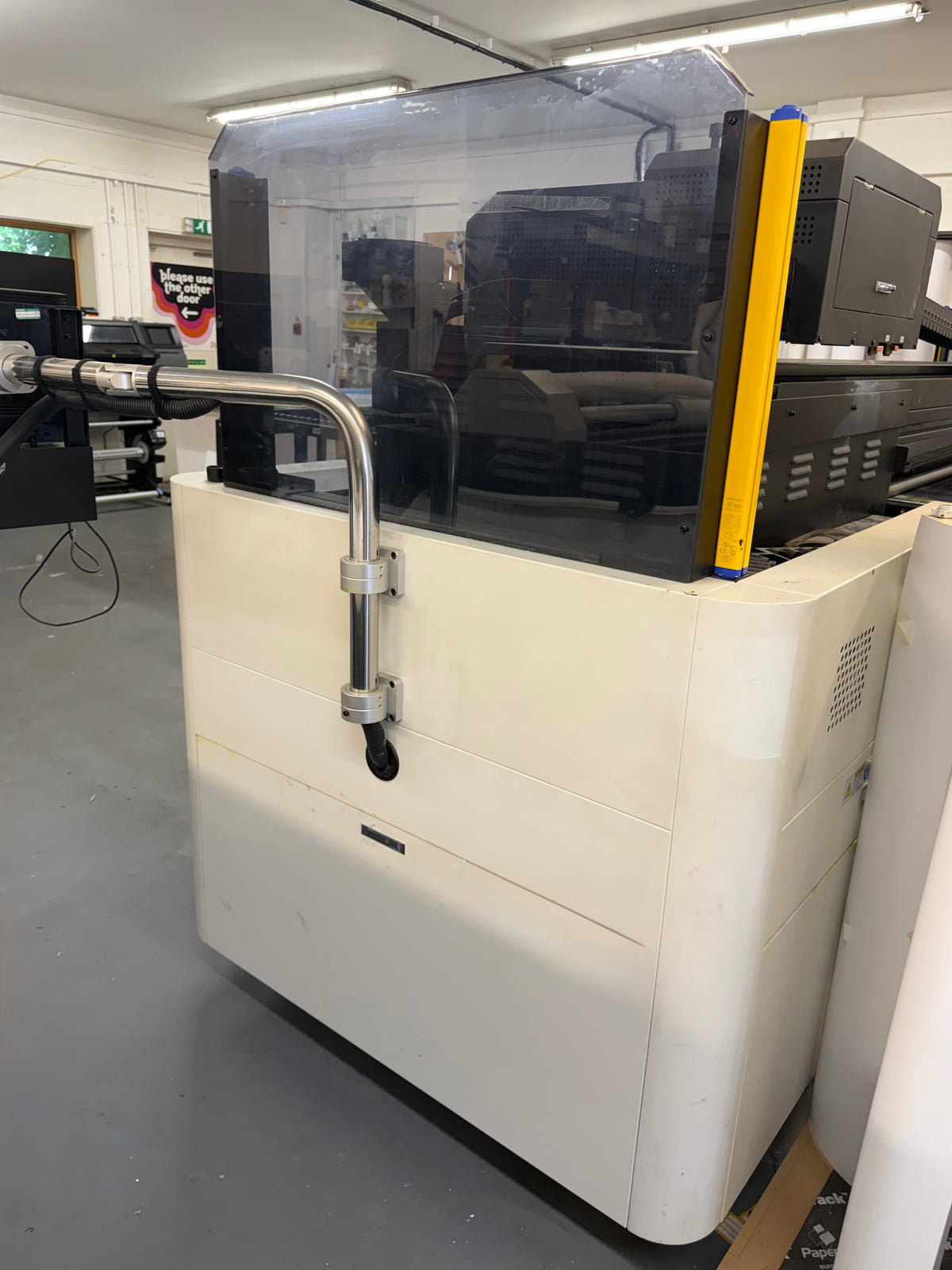 A large industrial printing machine with a cream-colored base, black upper housing, a clear protective cover, and a bright yellow vertical support arm, photographed in a workshop or studio interior.
