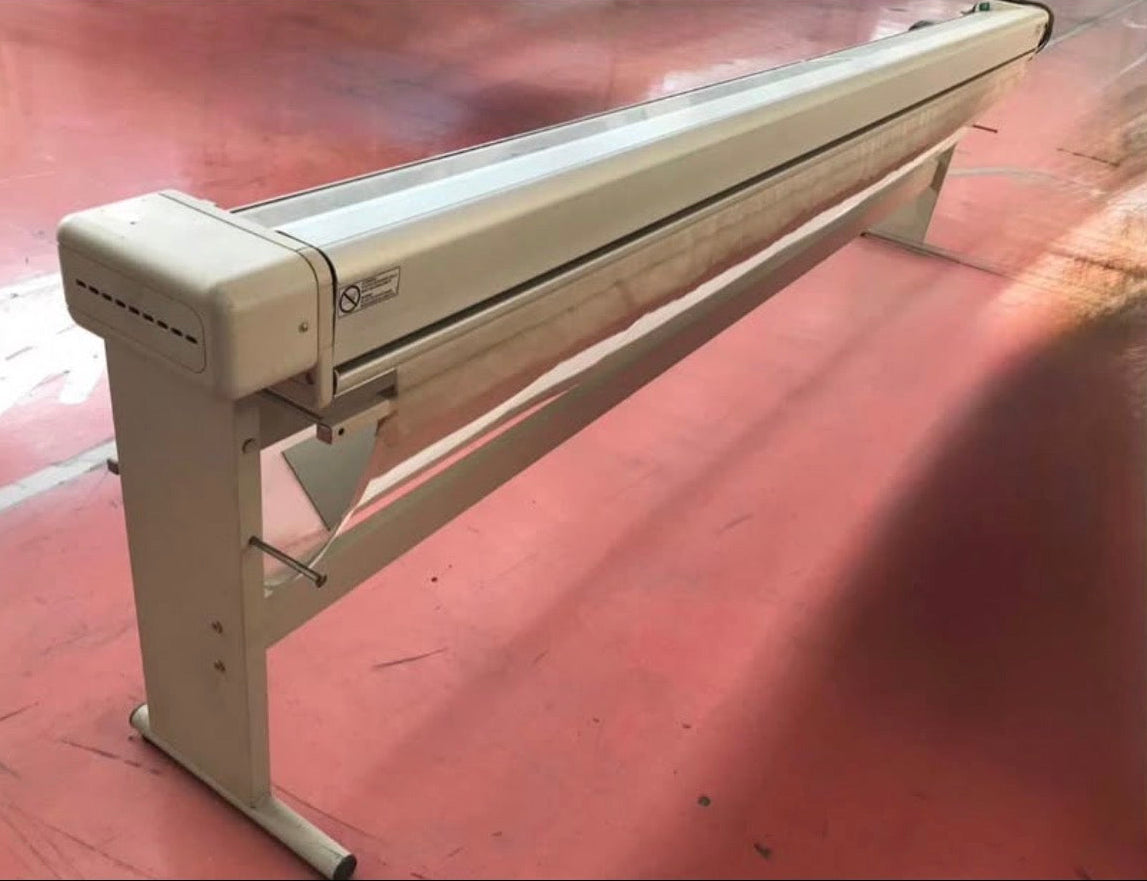 A long, narrow industrial machine or fixture with a beige frame and clear top panel, photographed on a dusty pink-red floor.