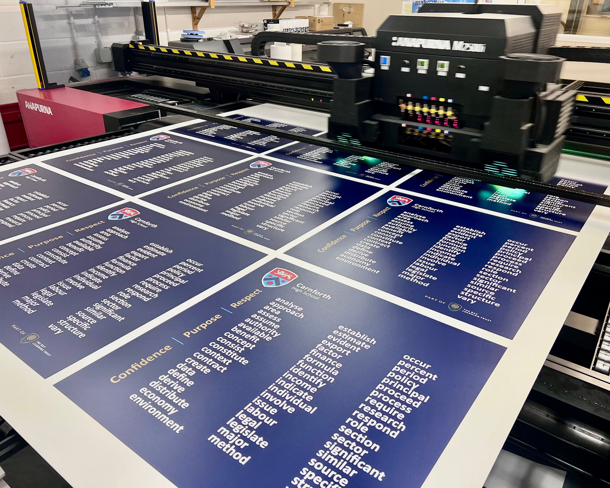A large printed board or poster with multiple blue panels and white text is being printed on by a large industrial UV printer in a factory setting. The board features the United States Air Force logo and text including 'Confidence', 'Principle', and 'Belief'.