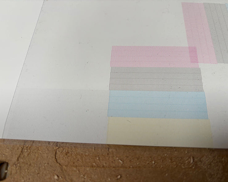 A close-up of a white sheet of paper with a faint pastel geometric pattern of light blue, pink, and gray blocks, positioned next to a brown textured surface.
